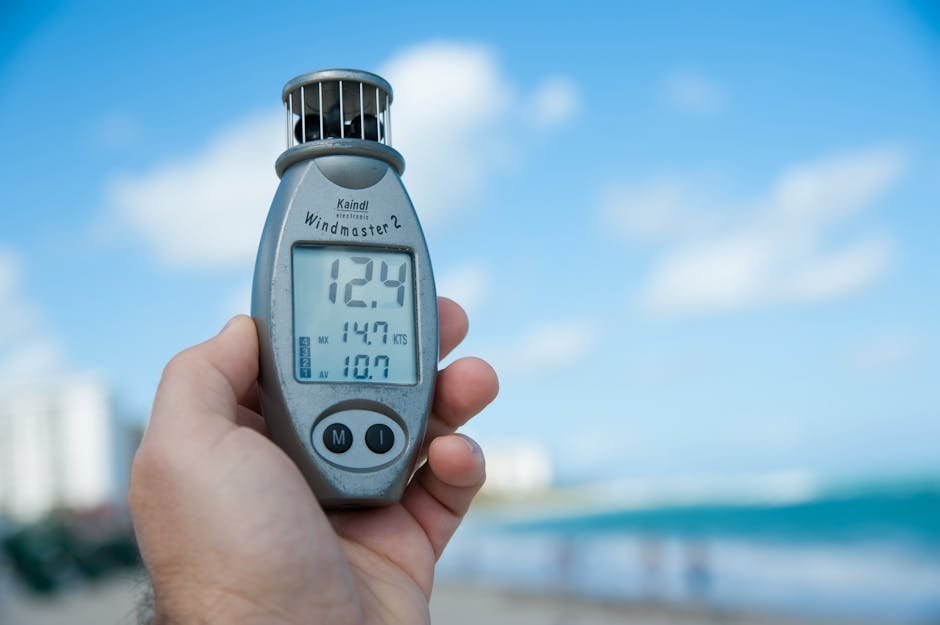 Close-up of a hand holding Windmaster 2 anemometer against a clear blue sky and beach backdrop.